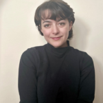 A photo of a person with short brown hair against a white backdrop wearing a black sweater.  The person is smiling in the photo. The photo is of Emma Baldwin.