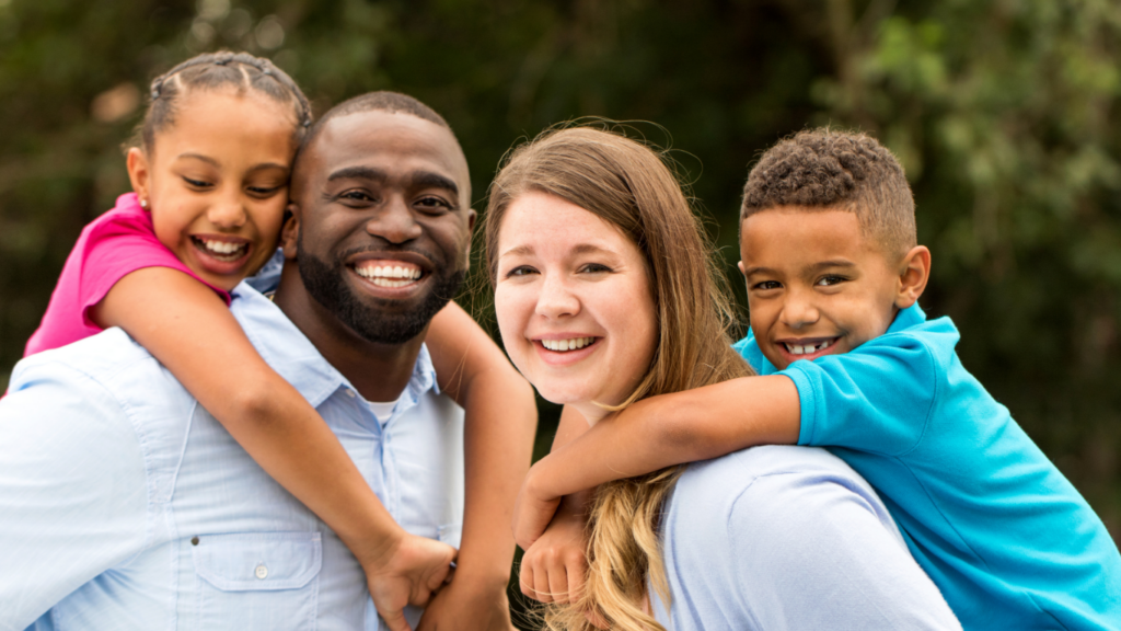 A family with two parents smile while holding their two kids on their backs.
