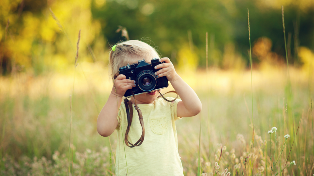 A young girl stands in a field holding a camera.